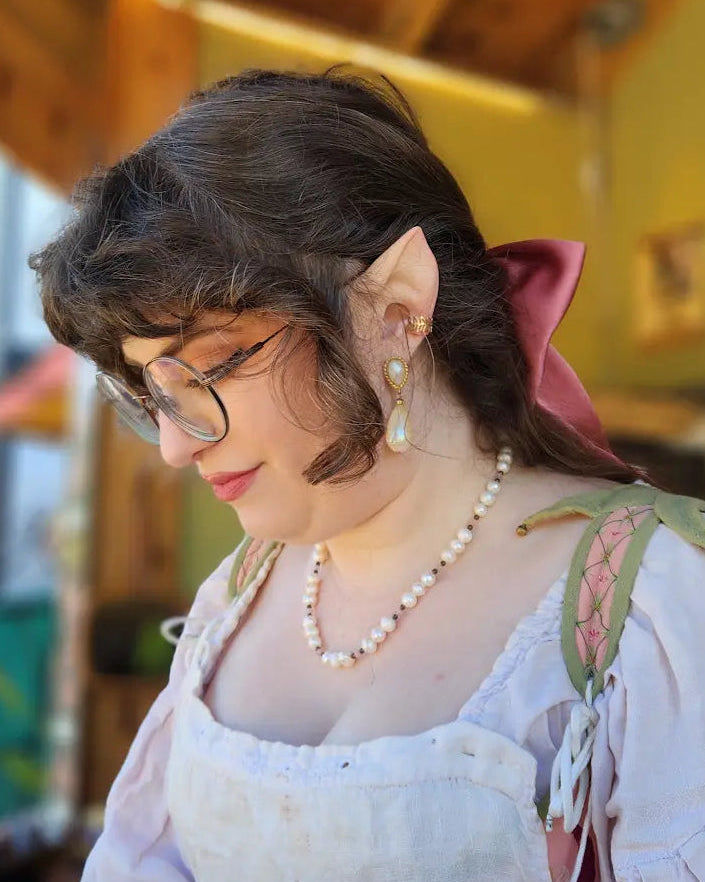 A woman wearing a medieval-style costume with a white, pink and green dress, pearl necklace, and glasses, looking down in a rustic indoor setting with wooden beams.