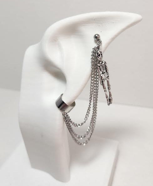 A silver-toned earring with multiple dangling chains and skull charms, displayed on a white plastic ear stand against a light gray background.