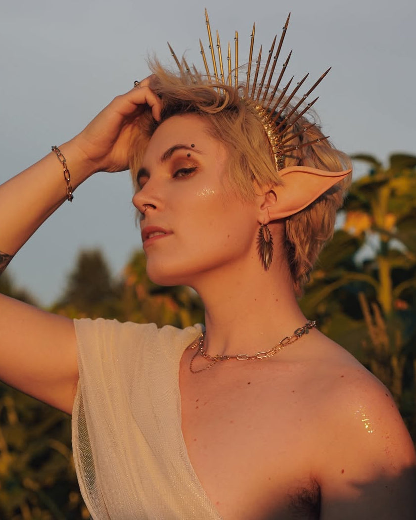 A person with blonde hair, pointed ears, and tattoos poses outdoors in a sunflower field at sunset, wearing a draped white toga-style dress, a gold crown, and a gold chain belt.
