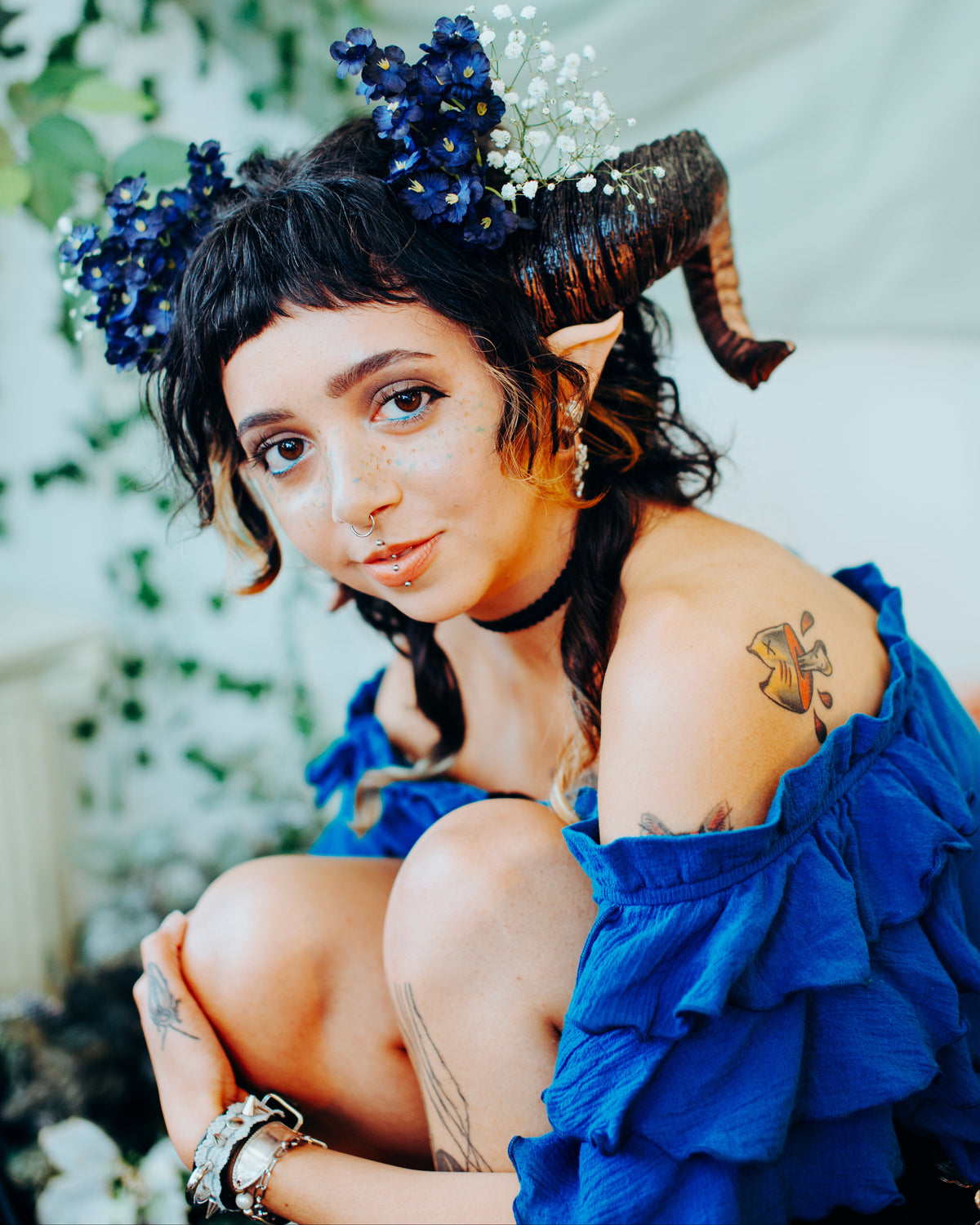 A woman in a fantasy costume wearing a bright blue ruffled dress, a horned headpiece, and a blue floral crown, seated against a soft draped backdrop with greenery.