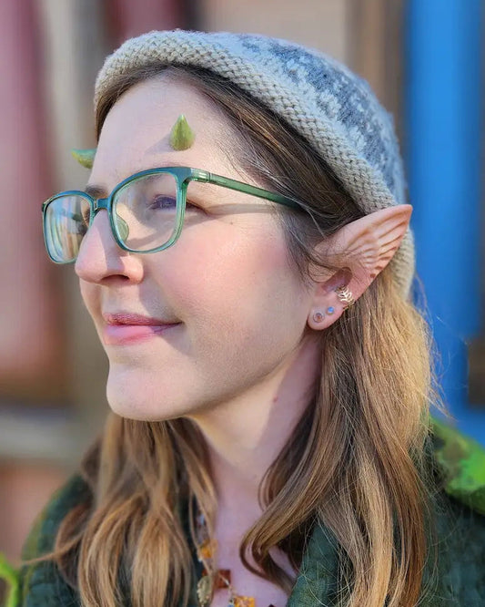 A woman with long light brown hair, wearing a gray knit elf hat, green glasses, and a textured dark green leafy costume jacket, shown in profile against a colorful blurred background.