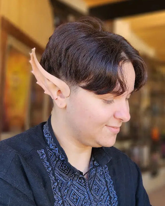 A person seated indoors wearing a pointed elf ear accessory on their ear, with a dark patterned top and a yellow 'Elf Ears' sign visible in the background.