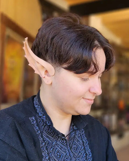A person seated indoors wearing a pointed elf ear accessory on their ear, with a dark patterned top and a yellow 'Elf Ears' sign visible in the background.
