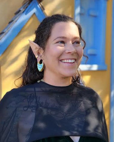 A smiling woman with pointed ears wearing fantasy elf ears, a black sheer cape, a dark green velvet top, and a gold patterned corset, standing outdoors in front of a yellow house with blue trim.
