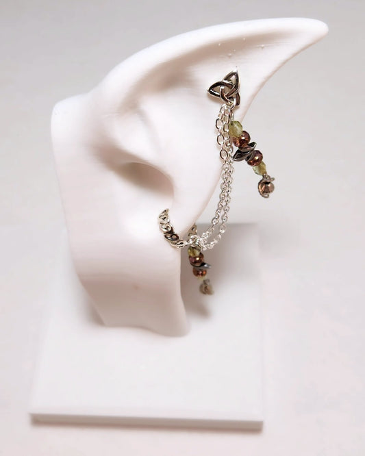 A silver-toned ear cuff with a Celtic knot design, chain links, and small green and brown beads, displayed on a white ear-shaped stand.