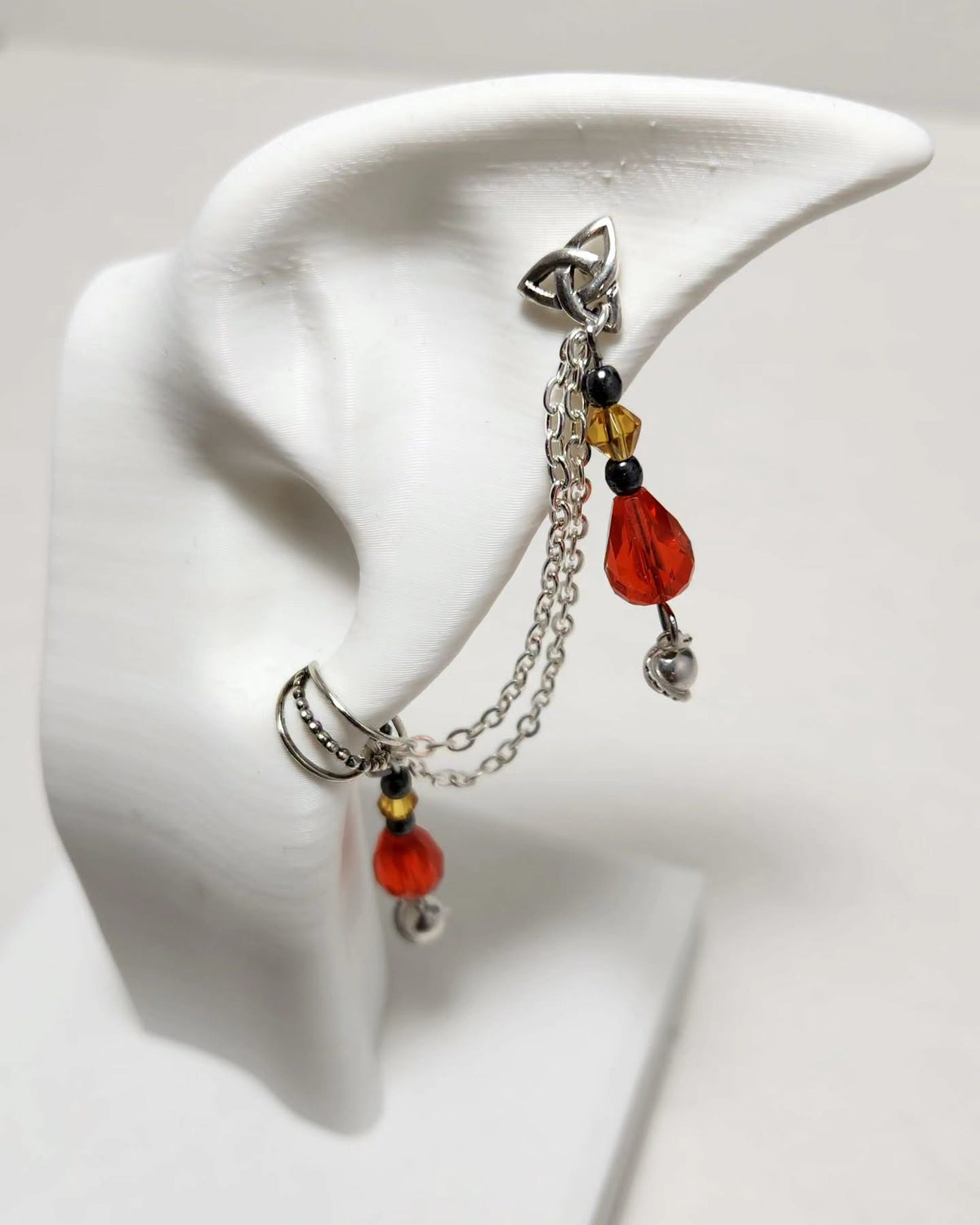 A silver-toned dangle earring displayed on a white ear-shaped jewelry stand. The earring features a triple-prong top, a short chain, a small amber bead, a translucent red teardrop bead, a black spacer bead, and a dangling silver bell.