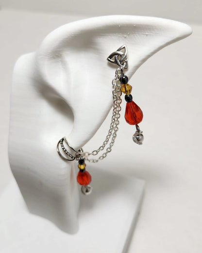 A silver-toned dangle earring displayed on a white ear-shaped jewelry stand. The earring features a triple-prong top, a short chain, a small amber bead, a translucent red teardrop bead, a black spacer bead, and a dangling silver bell.